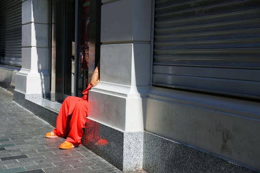 A person in bright clothing sits anonymously in a shadowed urban street corner.