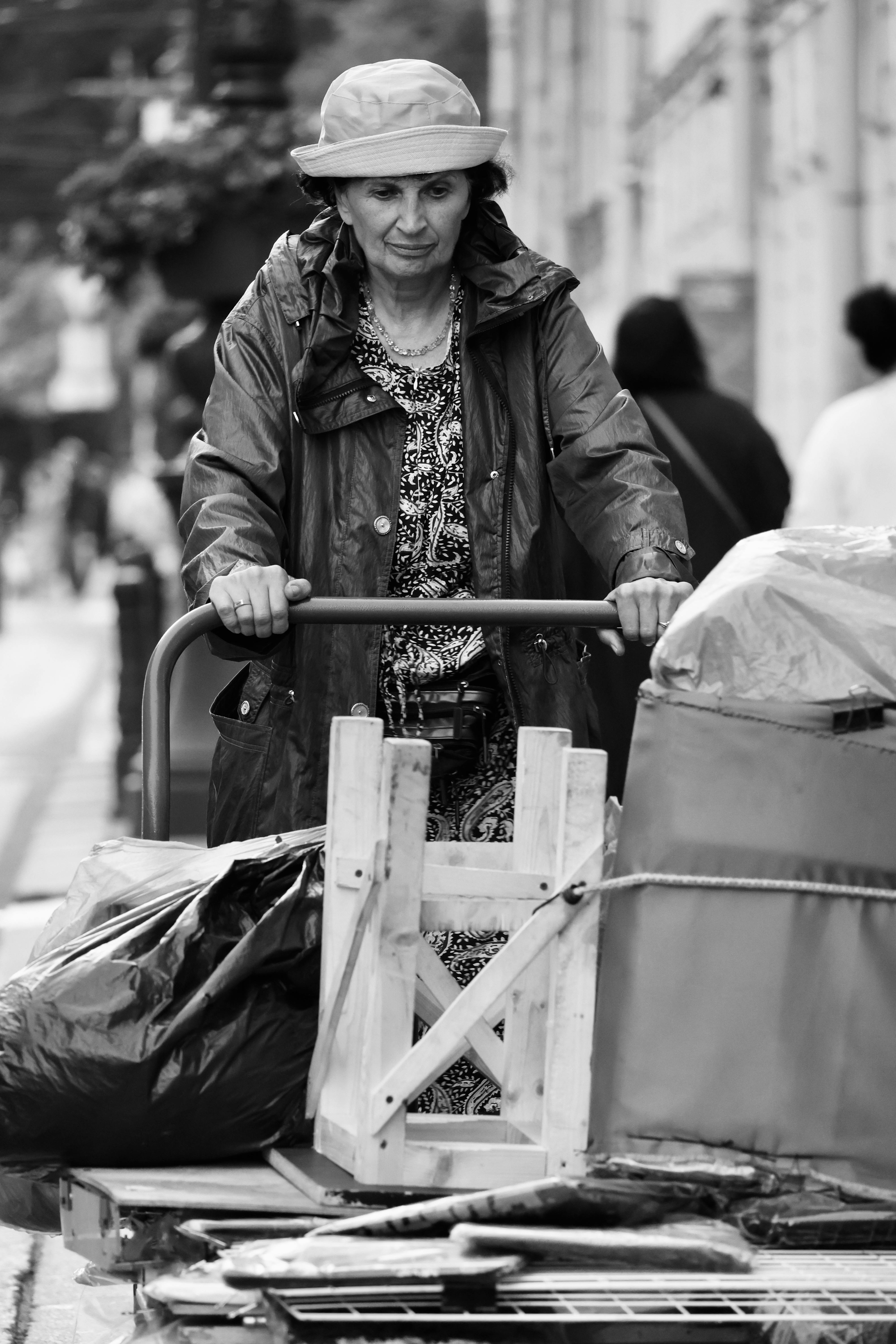 Black and white image of a woman pushing a cart filled with items on a rainy day street.