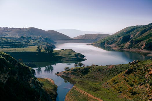 A stunning view of a tranquil lake surrounded by lush green hills and valleys under a clear blue sky.