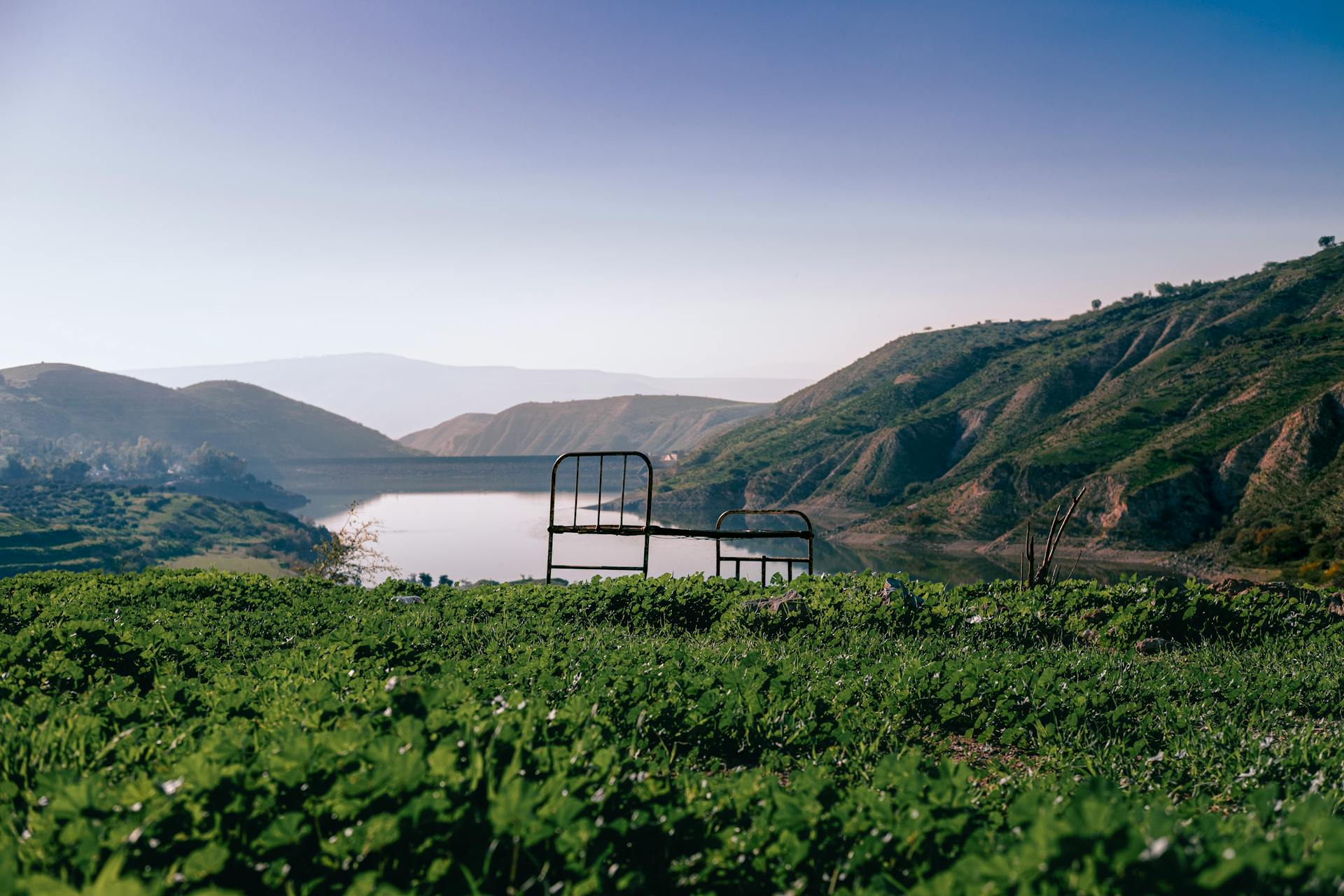 A serene lakeside view with an old bed frame amidst green hills and a clear blue sky.