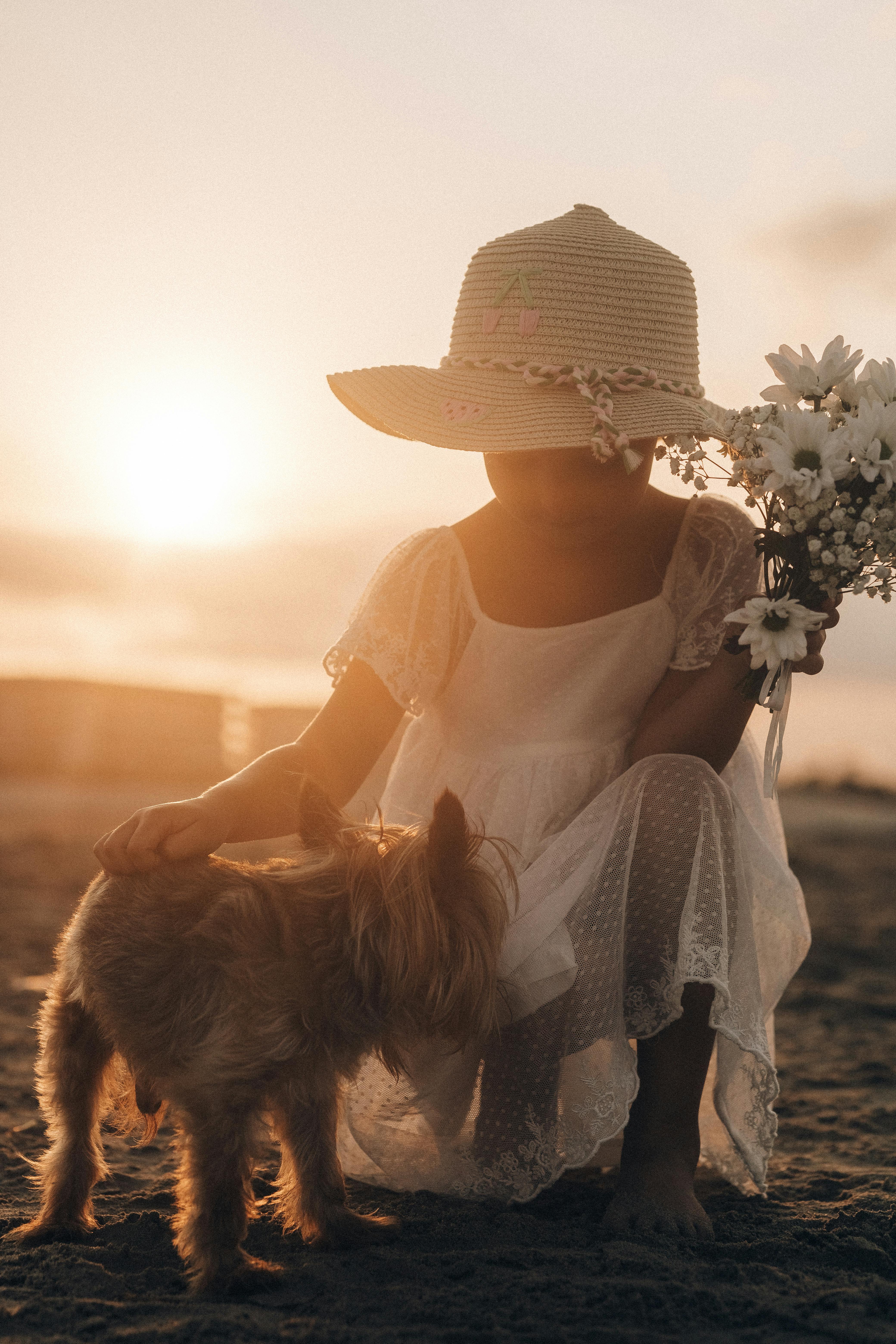 a little girl in a hat and dress petting a dog at sunset