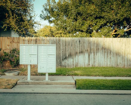 Mailboxes in a suburban area with a wooden fence and trees in the background.