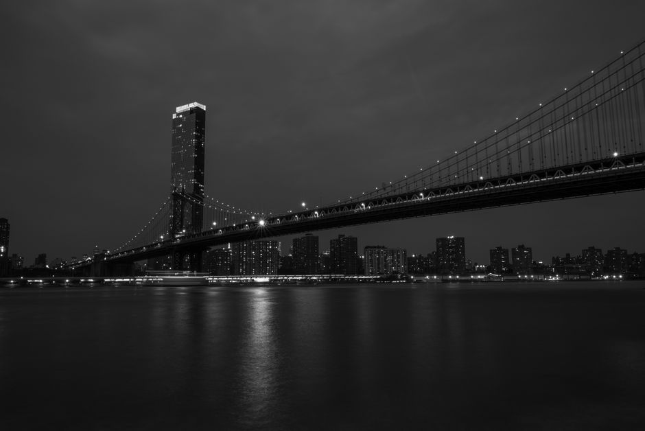 Dramatic black and white cityscape featuring a suspension bridge at night.