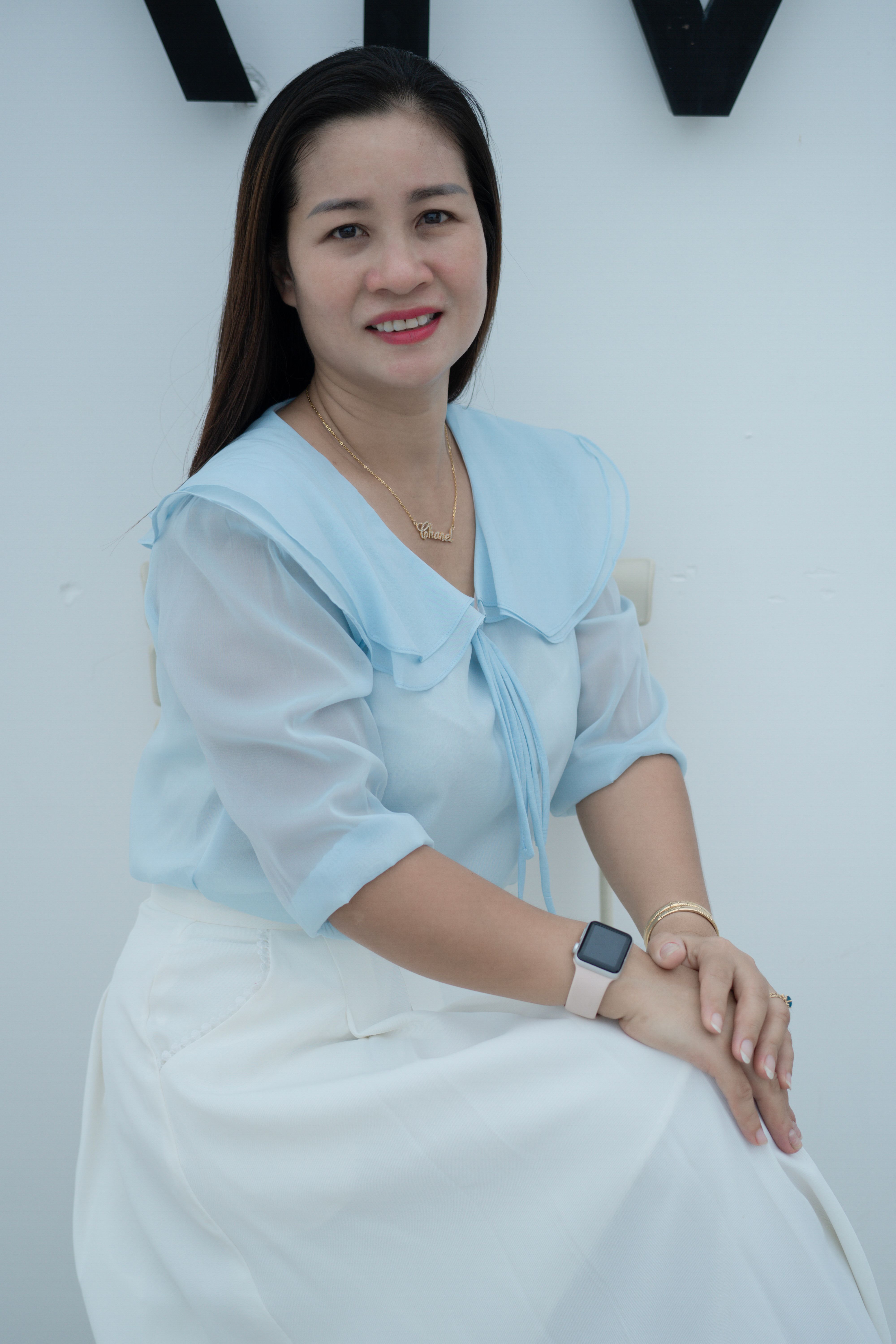A relaxed portrait of a smiling young woman sitting indoors, wearing a blue blouse and smartwatch.