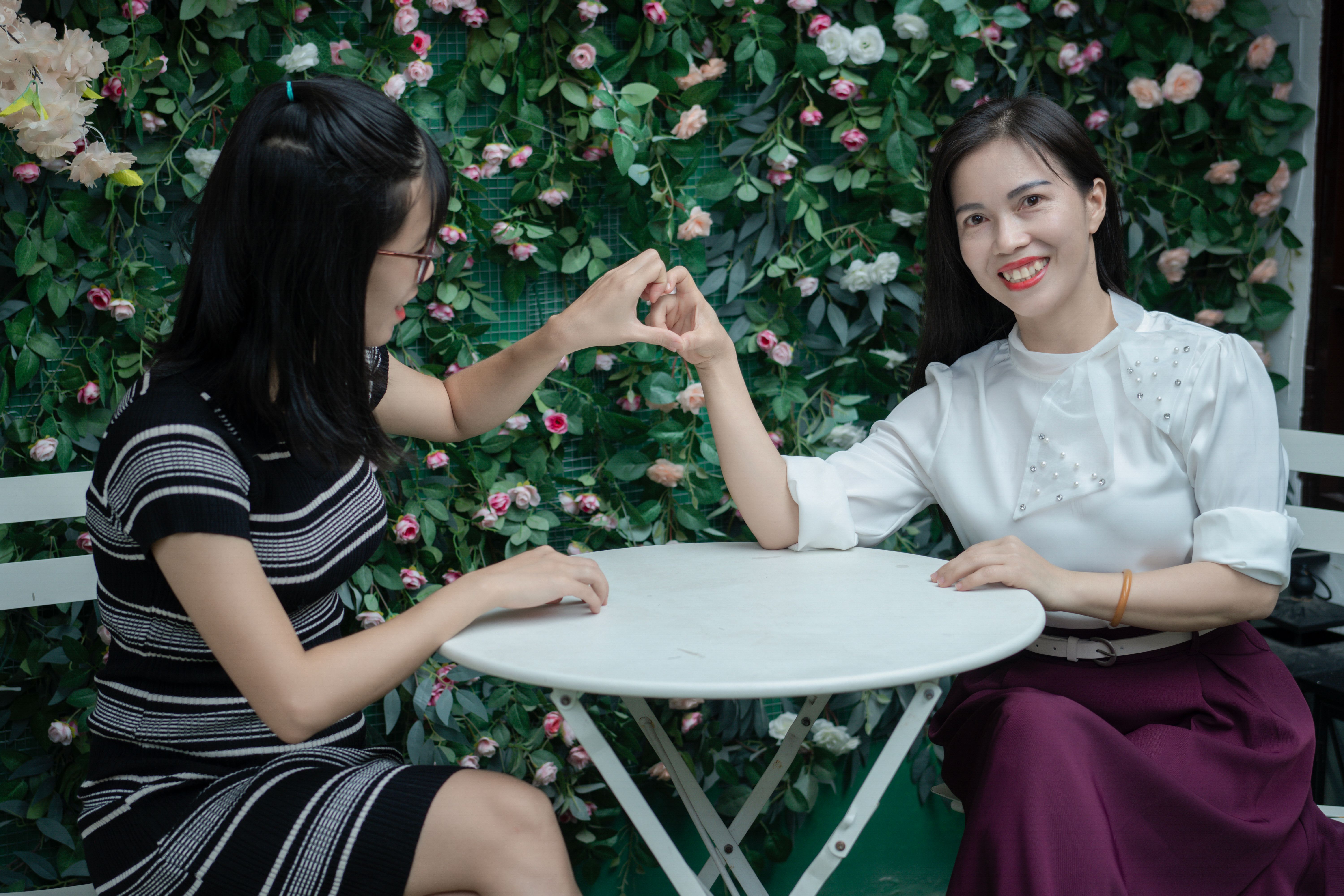 Two women interact joyfully at a cafe table surrounded by vibrant floral decor.