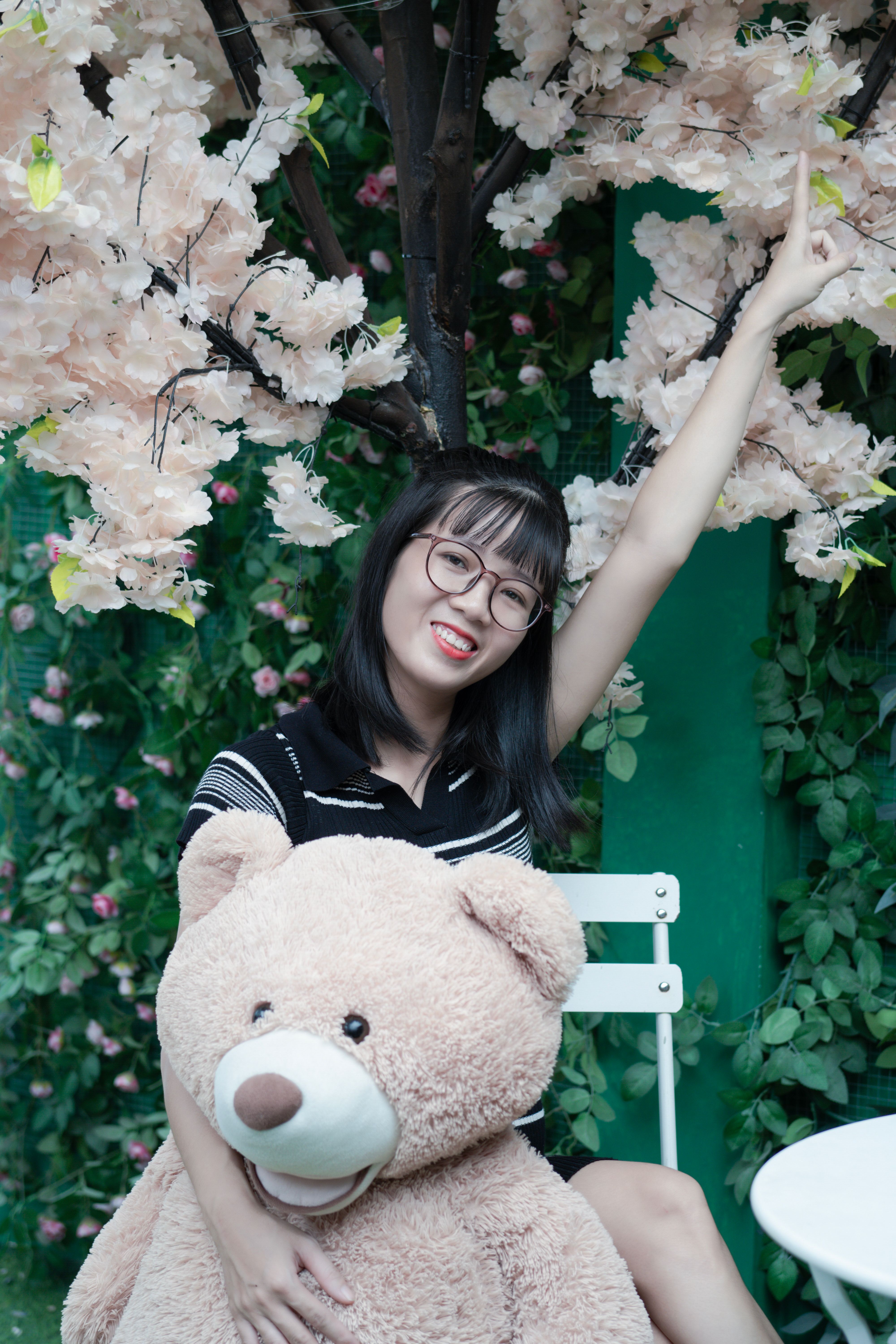 A joyful woman with glasses sits with a teddy bear under cherry blossoms outdoors, portraying happiness.