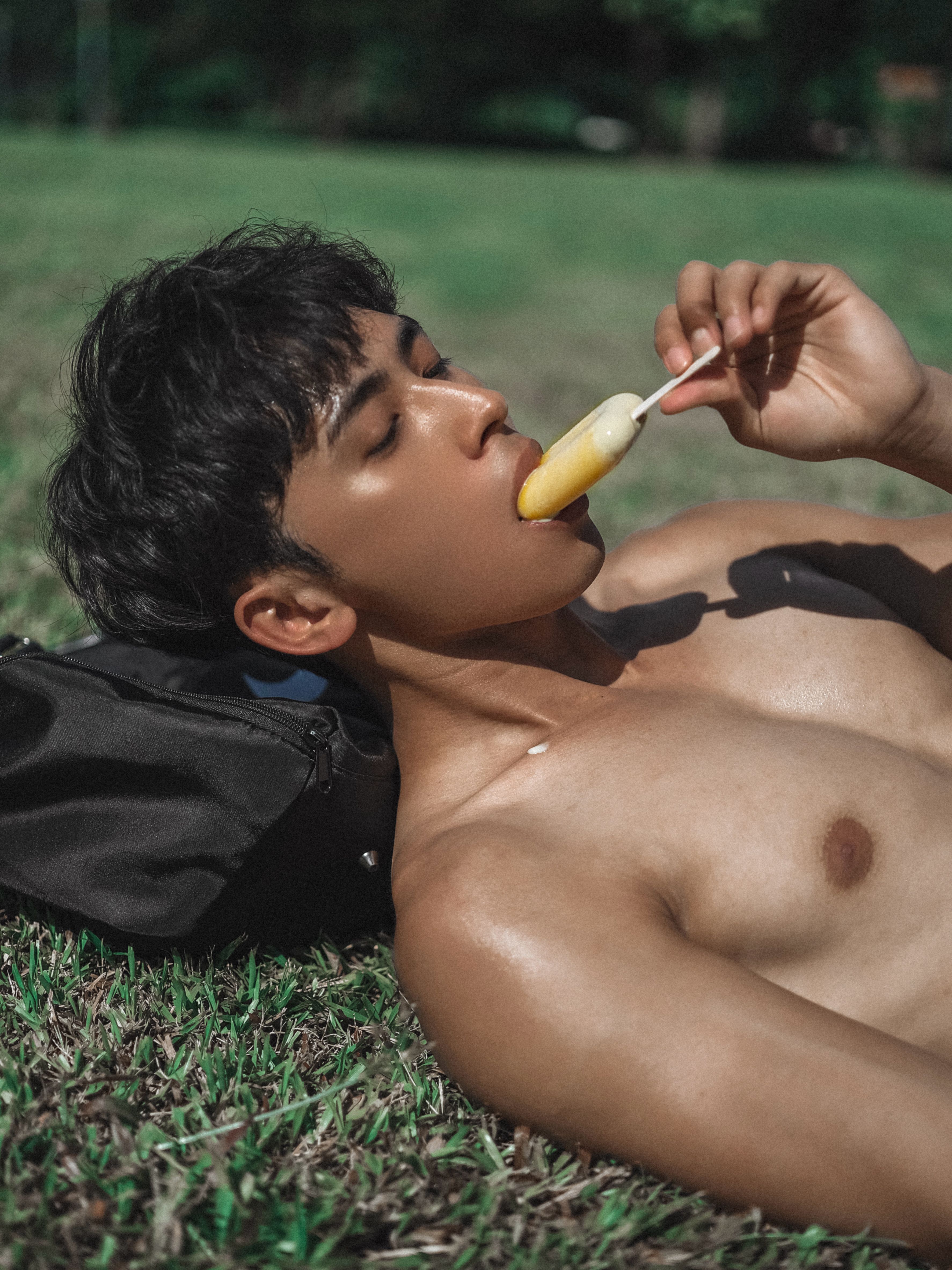 Shirtless young man enjoying a popsicle while reclining on grass in a sunny outdoor setting.