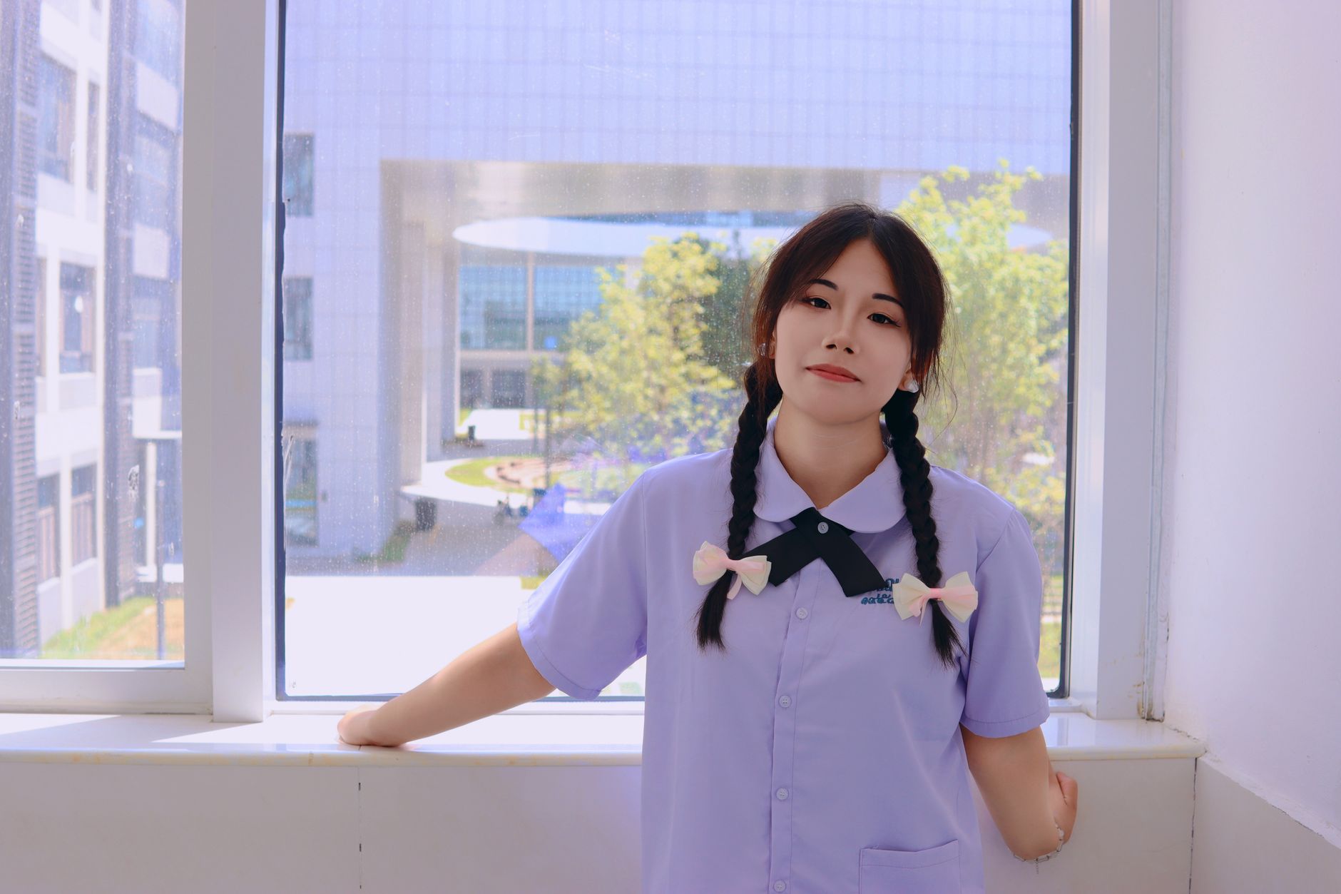 Young woman in a uniform stands by a window with a view of a modern building, exuding a calm demeanor.