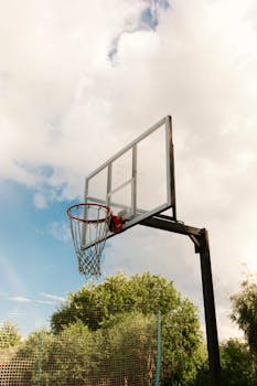 A basketball hoop with netting and clear backboard set outdoors under a cloudy blue sky.