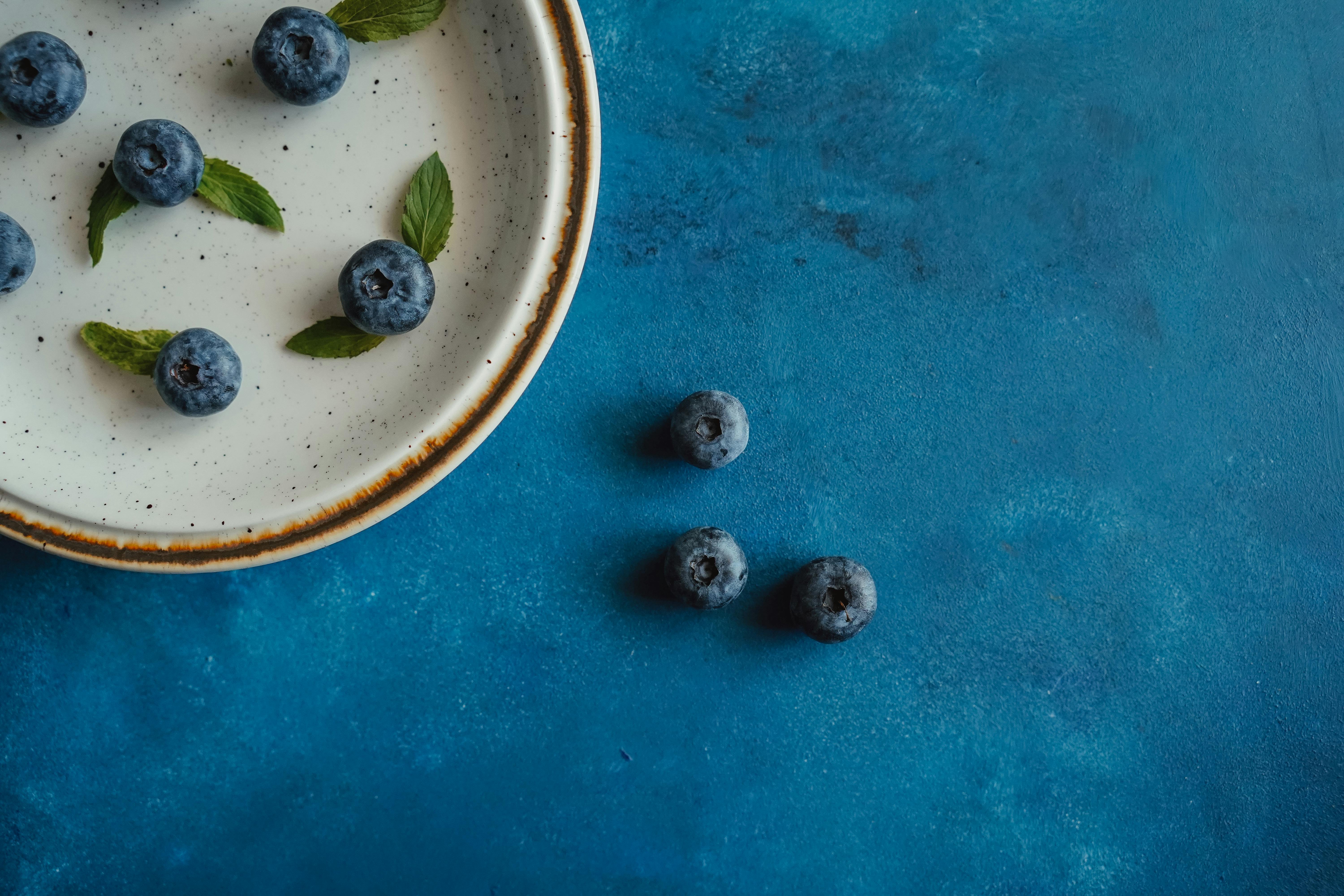 Overhead view of fresh blueberries in a rustic ceramic bowl with mint leaves on a blue surface.