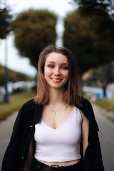 A cheerful young woman in a white top and black jacket smiling on a tree-lined street.