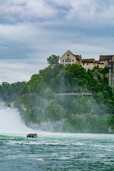 Historic castle overlooking the majestic Rhine Falls in Switzerland, with lush greenery and a boat on the water.
