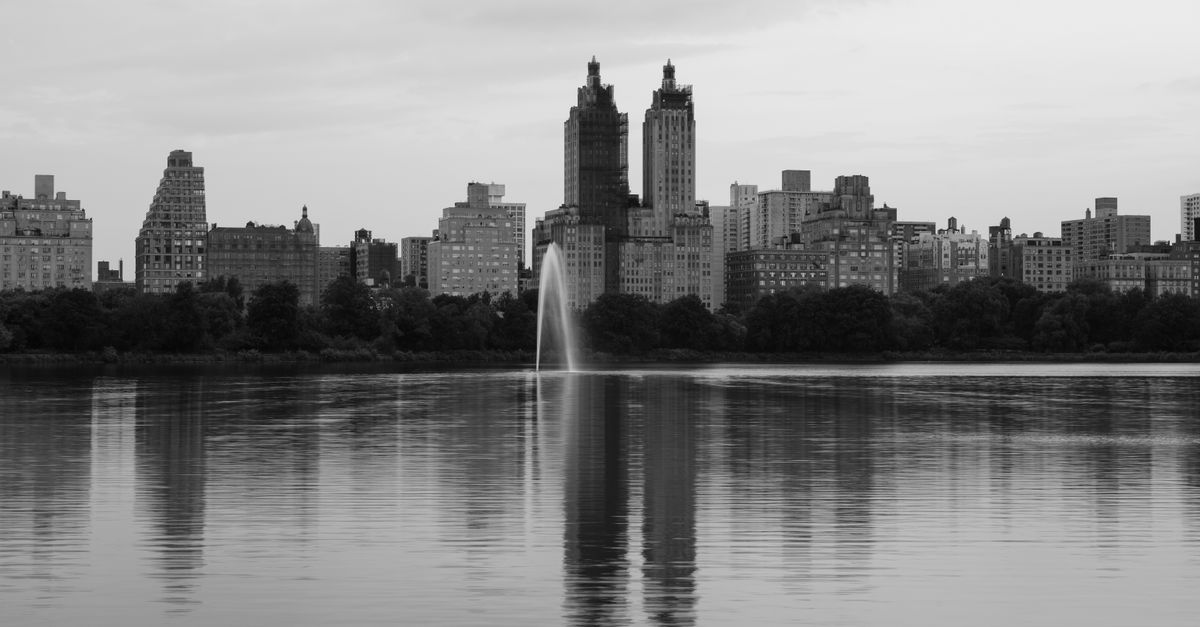 Black and white image of New York City's skyline reflecting in a calm lake with a visible water fountain.