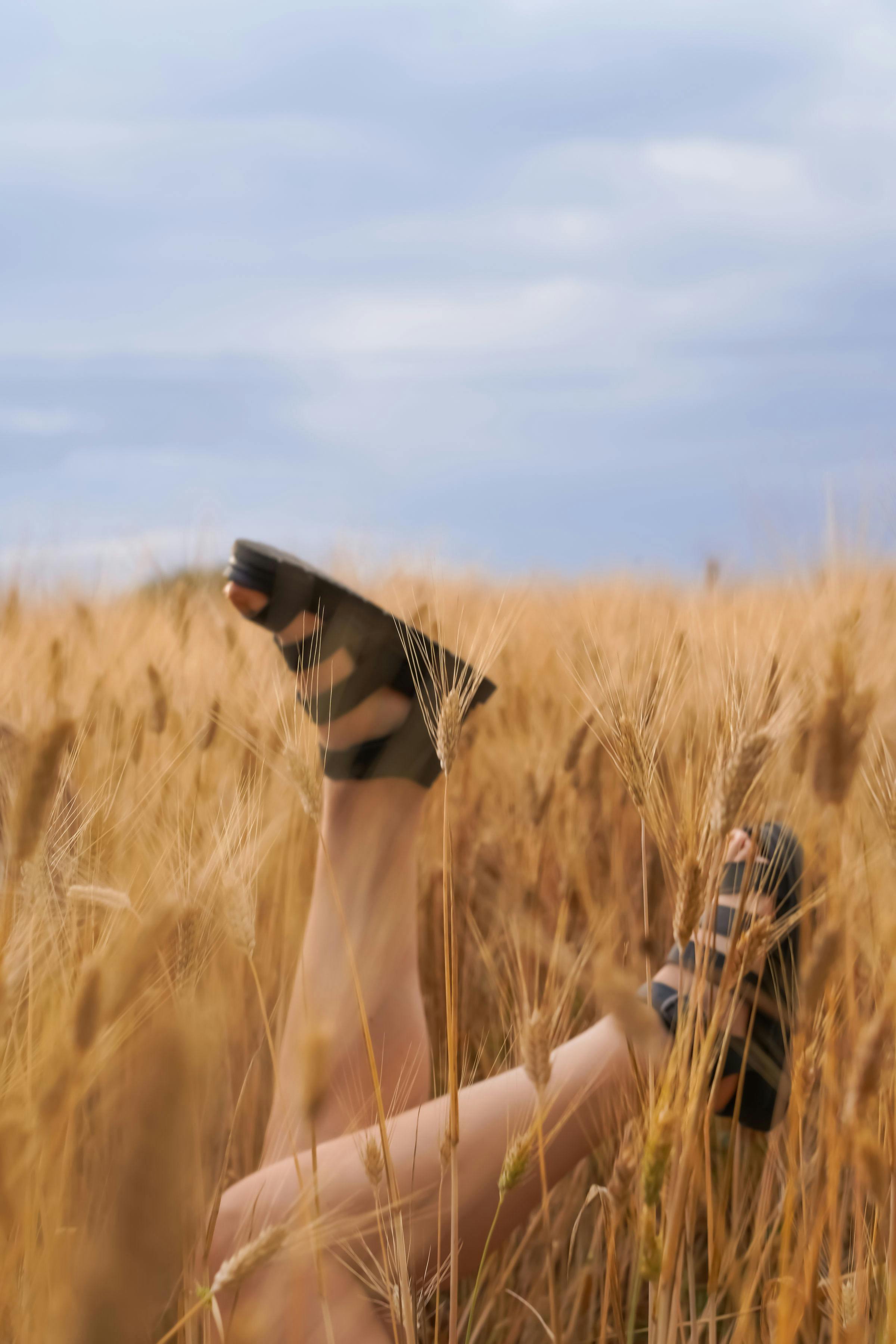 Relaxing in a wheat field with sandals visible among golden grains under a cloudy sky.