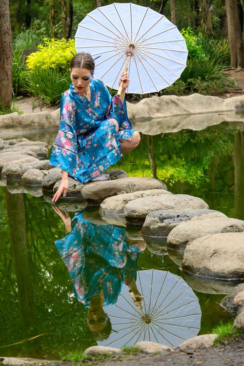 Free A serene portrait of a woman in a floral blue kimono holding an umbrella by a reflective pond. Stock Photo