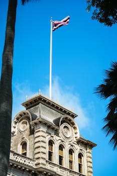 View of Iolani Palace in Honolulu featuring the Hawaiian flag against a clear blue sky.