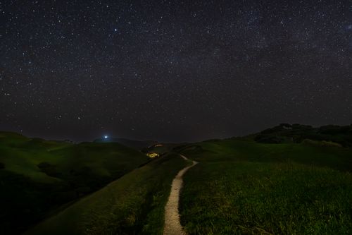 Free A clear starry night sky illuminates a winding path through lush grassy hills. Stock Photo