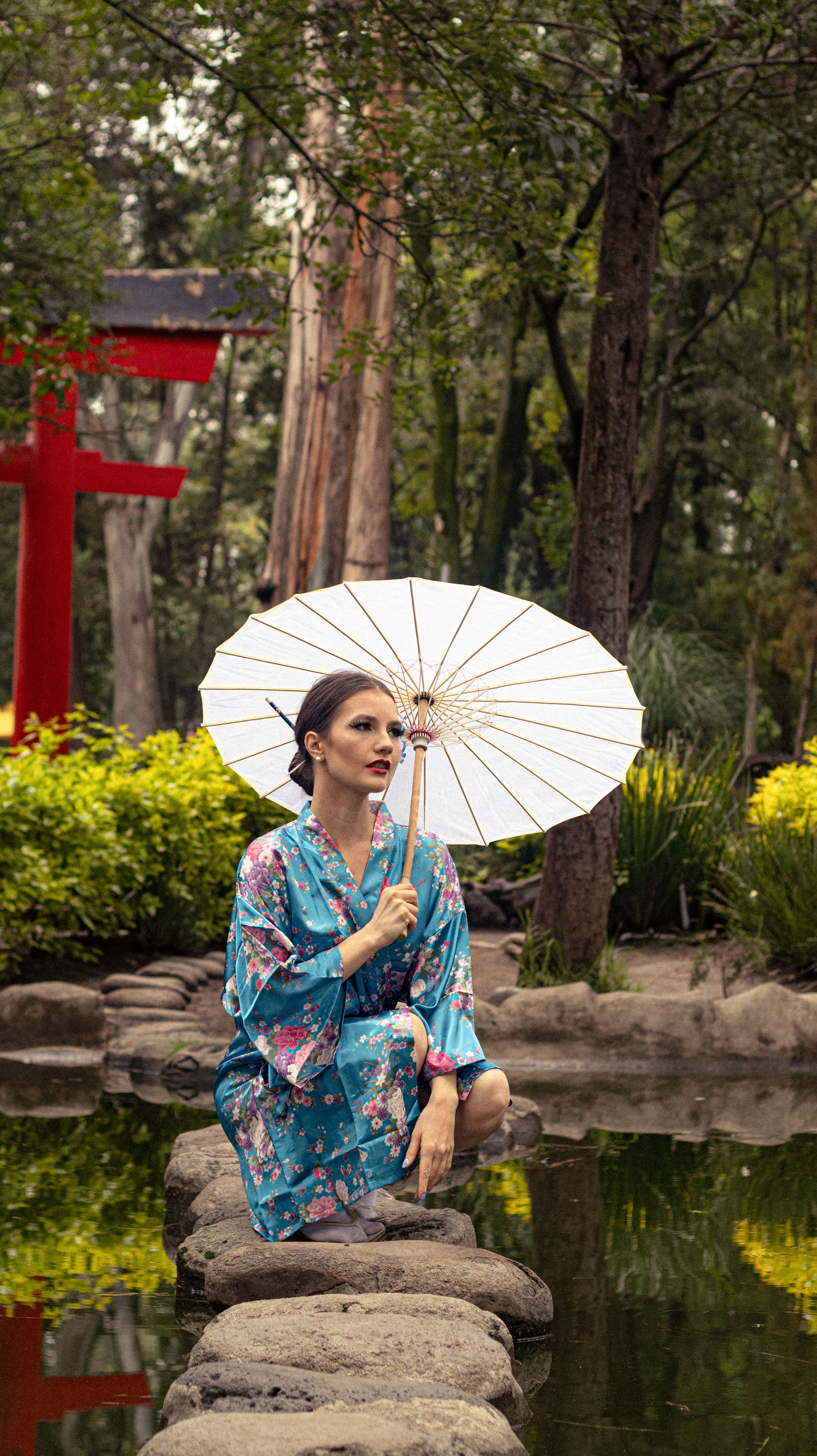 Woman in floral kimono with a white parasol by a tranquil pond in a Japanese garden.