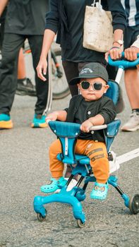 Cute toddler in sunglasses enjoying a ride on a blue tricycle in a lively street scene. - Fotografer Sidoarjo