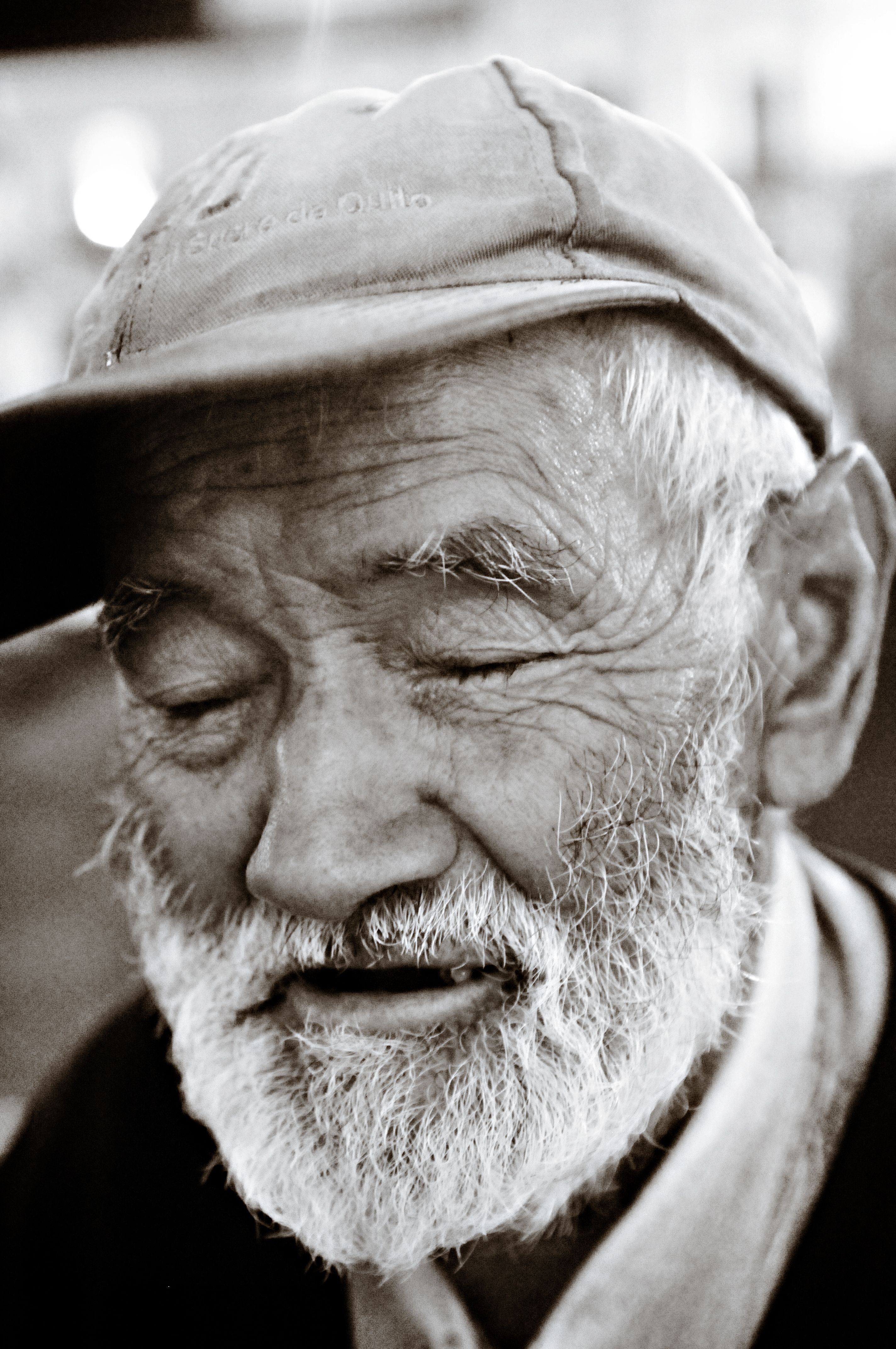 A candid black and white portrait of a smiling elderly man with a beard and cap, captured outdoors.