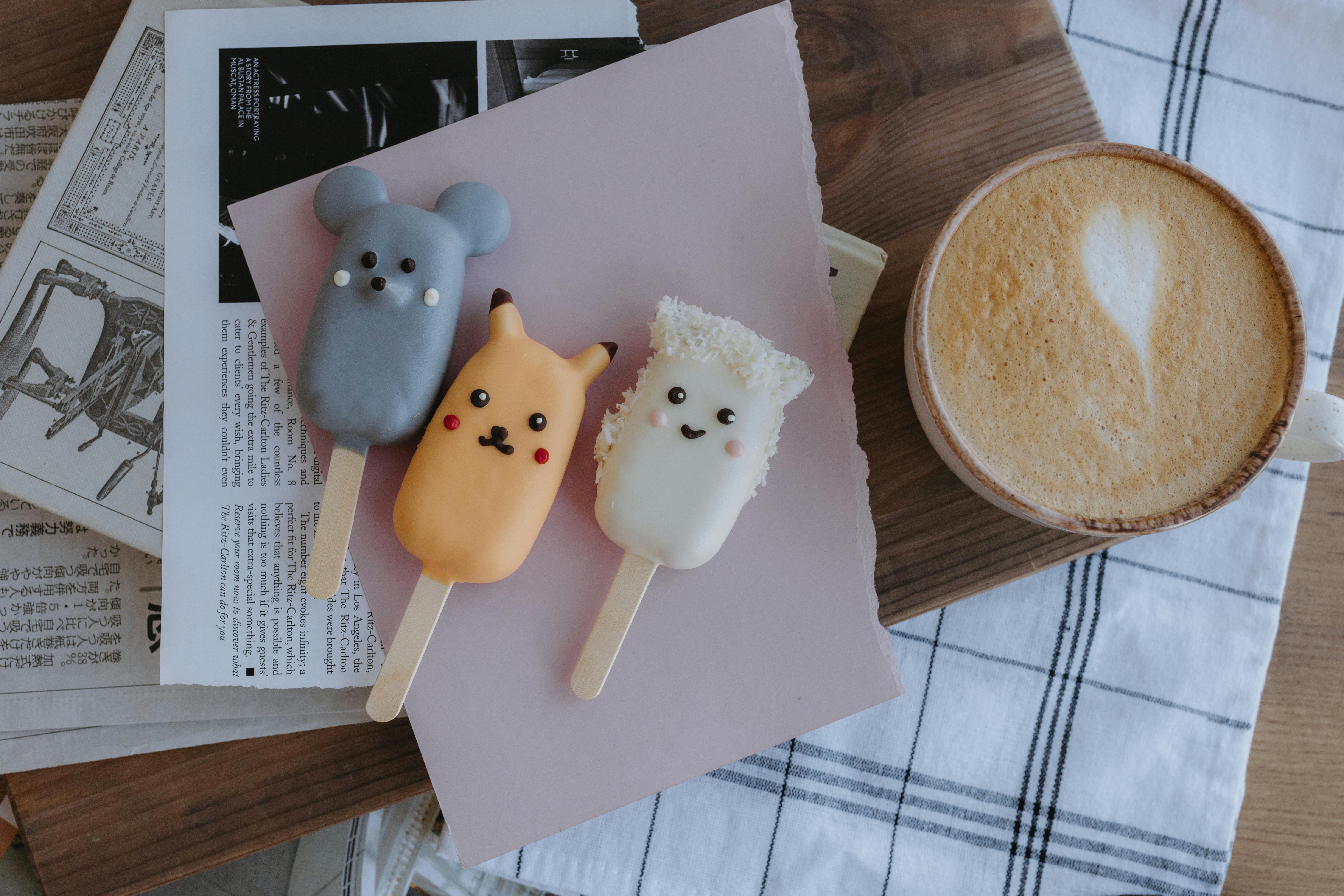 popsicles with cute animals on them sit on a table next to a cup of coffee