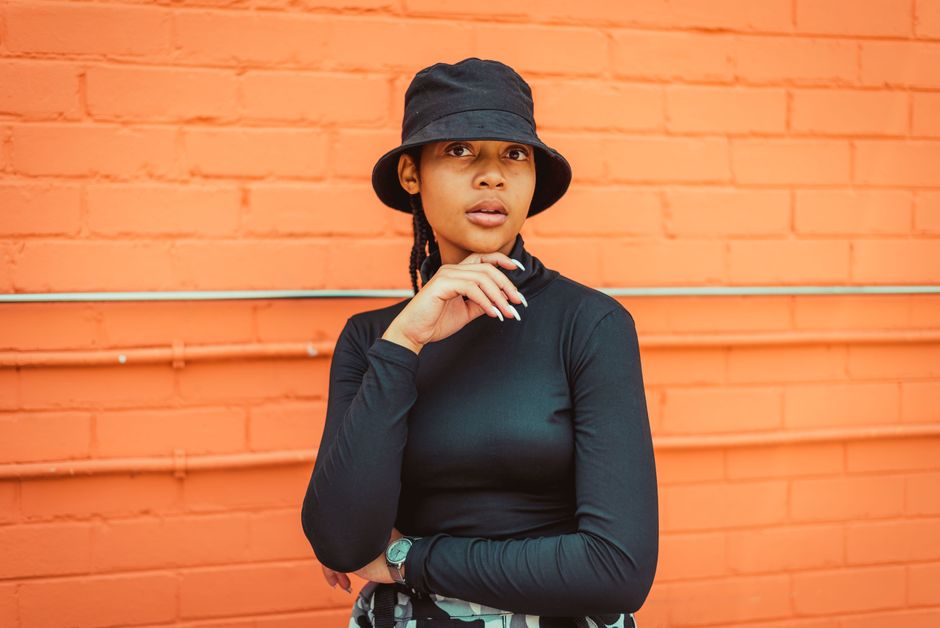 Stylish woman in a black hat and shirt poses confidently against an orange brick wall, showcasing modern street fashion.