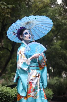 Woman in a blue kimono holding an umbrella and fan, outdoors in a lush setting.