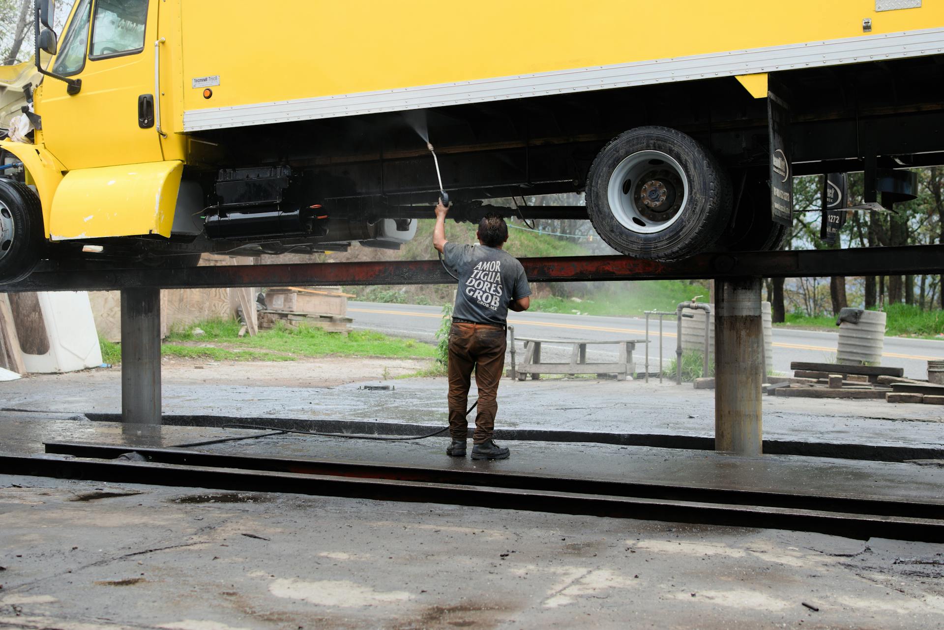 A worker power washes the underside of a raised yellow truck at an outdoor service station.