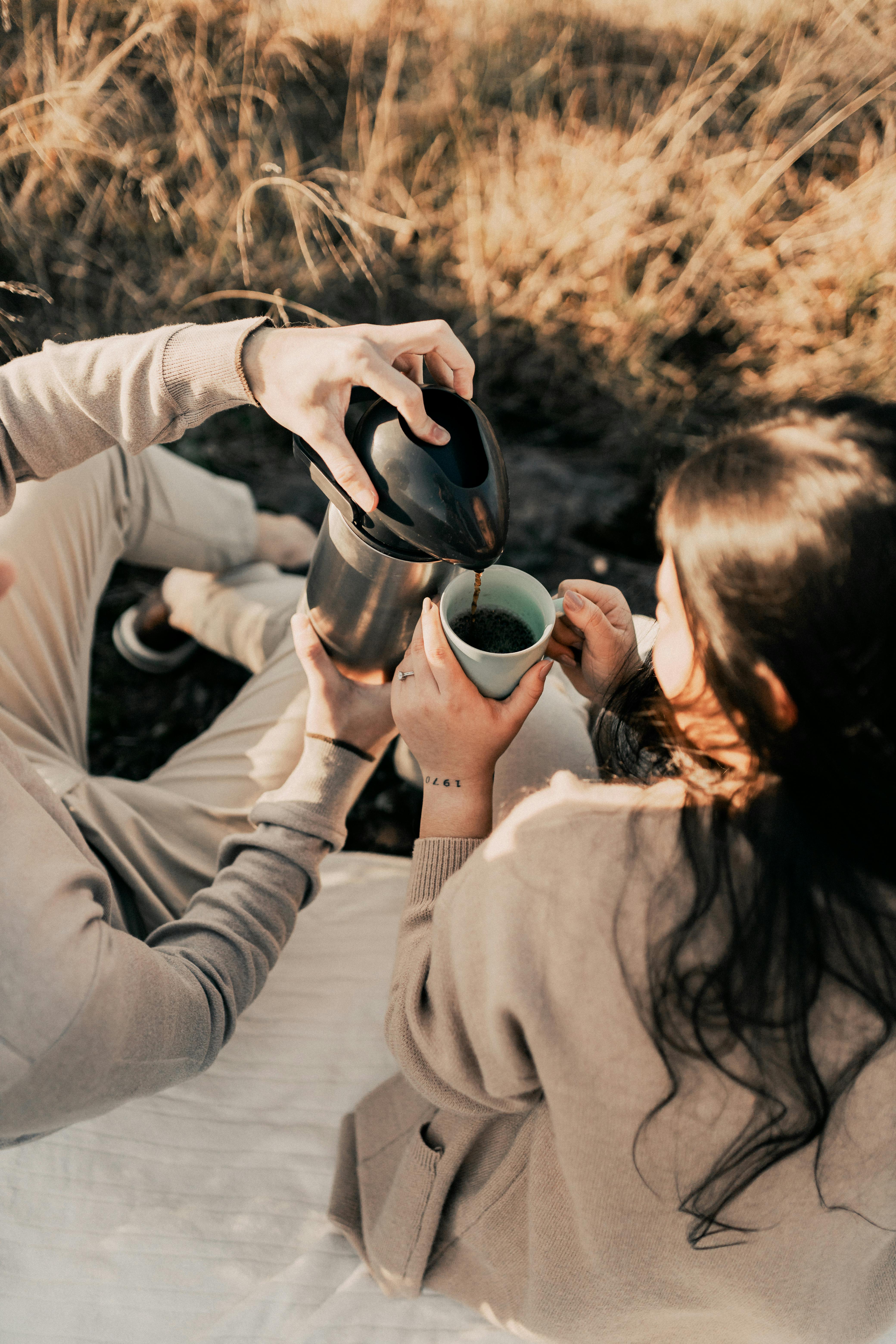 A couple enjoys a warm coffee together in a sunlit field during a summer day.