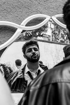 Black and white photo of a man in a leather jacket smoking, with a mirror reflection outdoors.