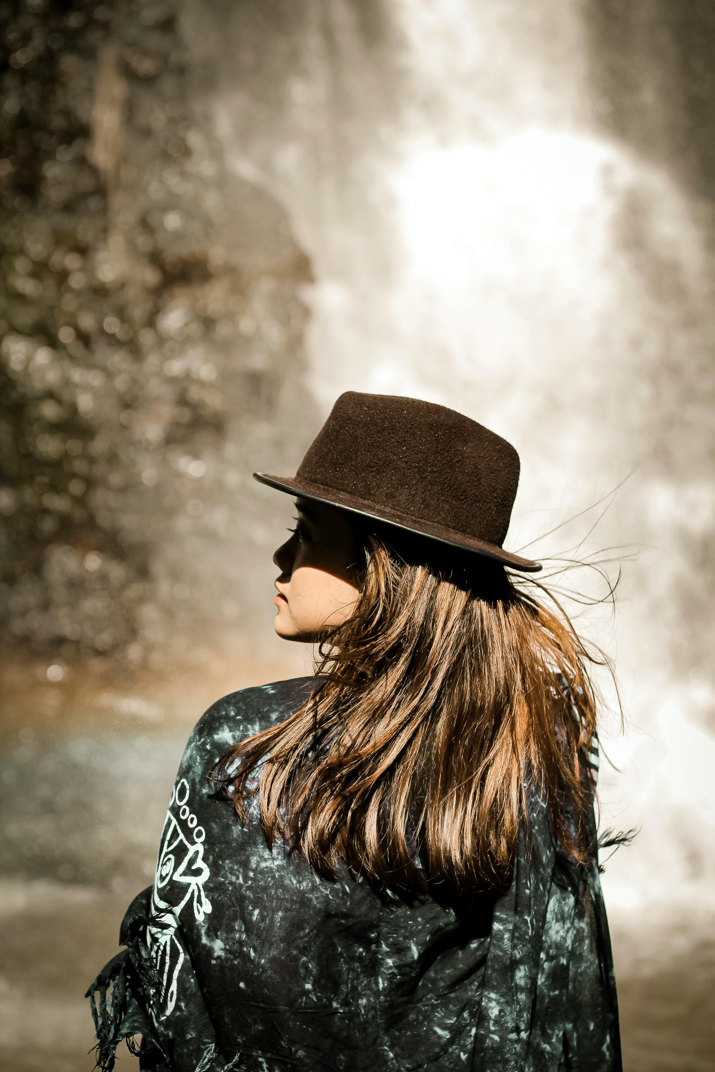 一位年轻女子戴着帽子，站在风景如画的瀑布旁，感受着大自然的宁静气息