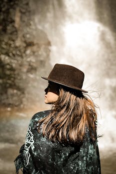 一位年轻女子戴着帽子，站在风景如画的瀑布旁，感受着大自然的宁静气息