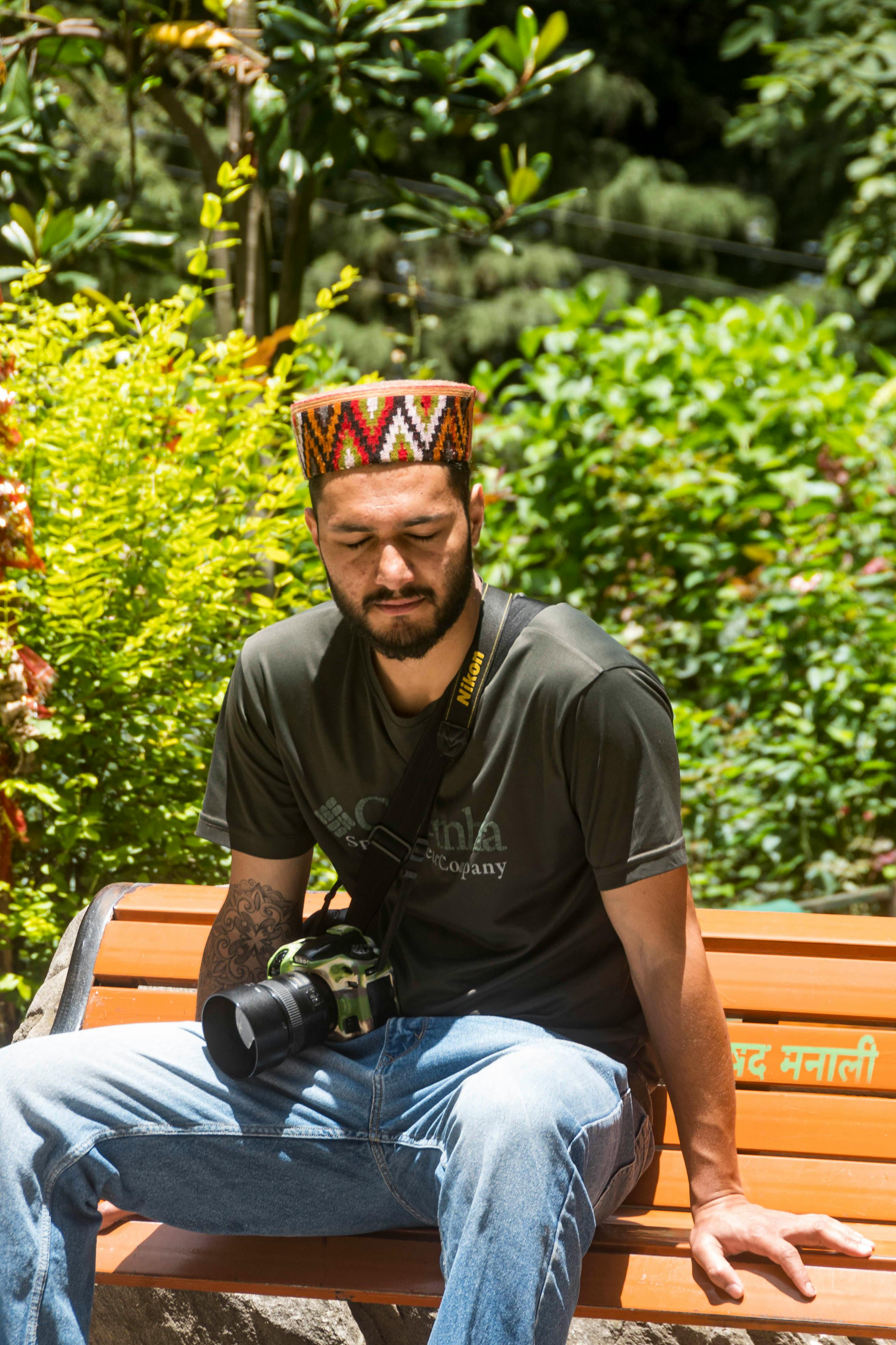 A man with a colorful cap relaxes on a bench in a sunny, lush garden.