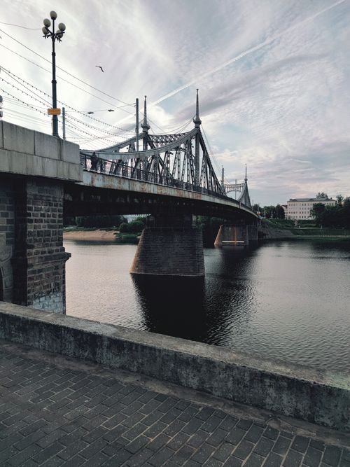 Free Historic steel bridge over a calm river, offering urban landscape views with cloudy sky reflections. Stock Photo