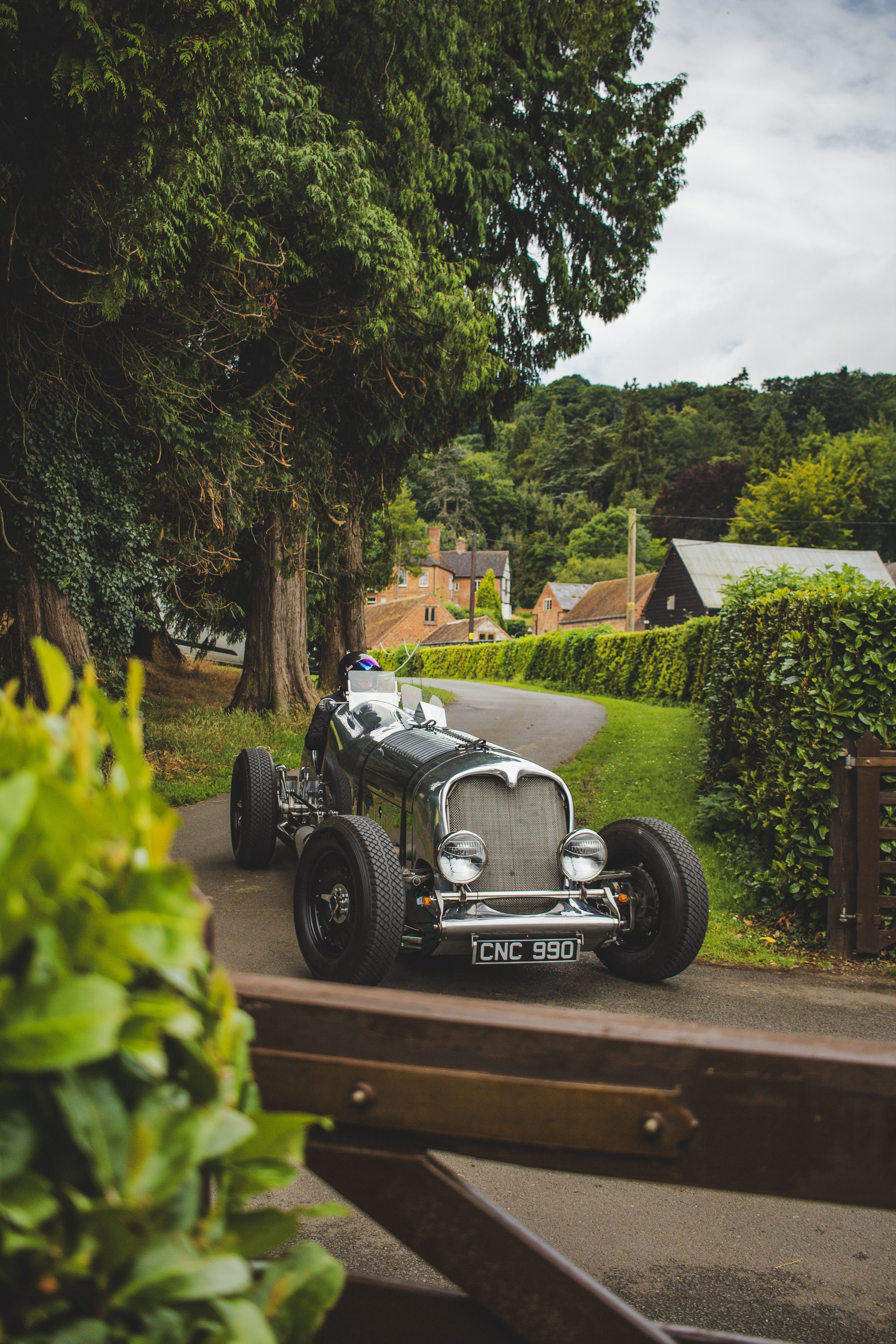 A vintage racing car driving on a picturesque country road surrounded by lush greenery.