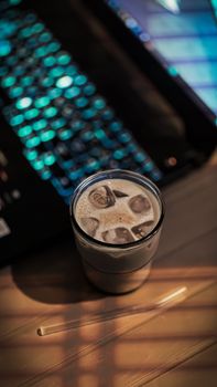 A refreshing glass of iced coffee on a wooden desk by a glowing laptop keyboard, creating a cozy evening ambiance.
