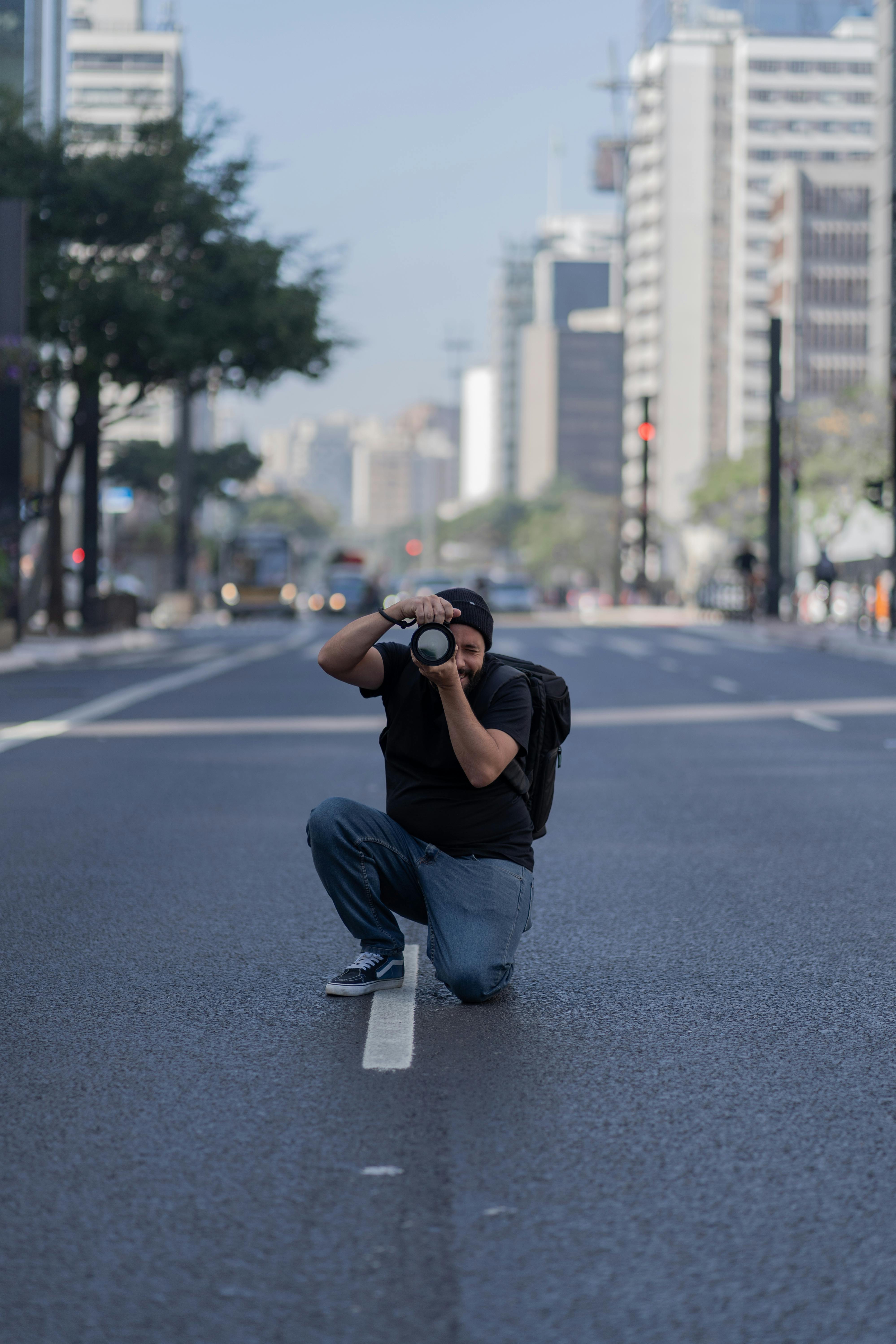A photographer kneeling beside a puddle, holding a camera with a macro lens, with Auckland city skyline in the background.