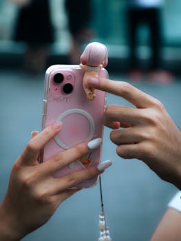 Hands holding a pink iPhone with a toy charm in Bangkok, Thailand, captured outdoors.