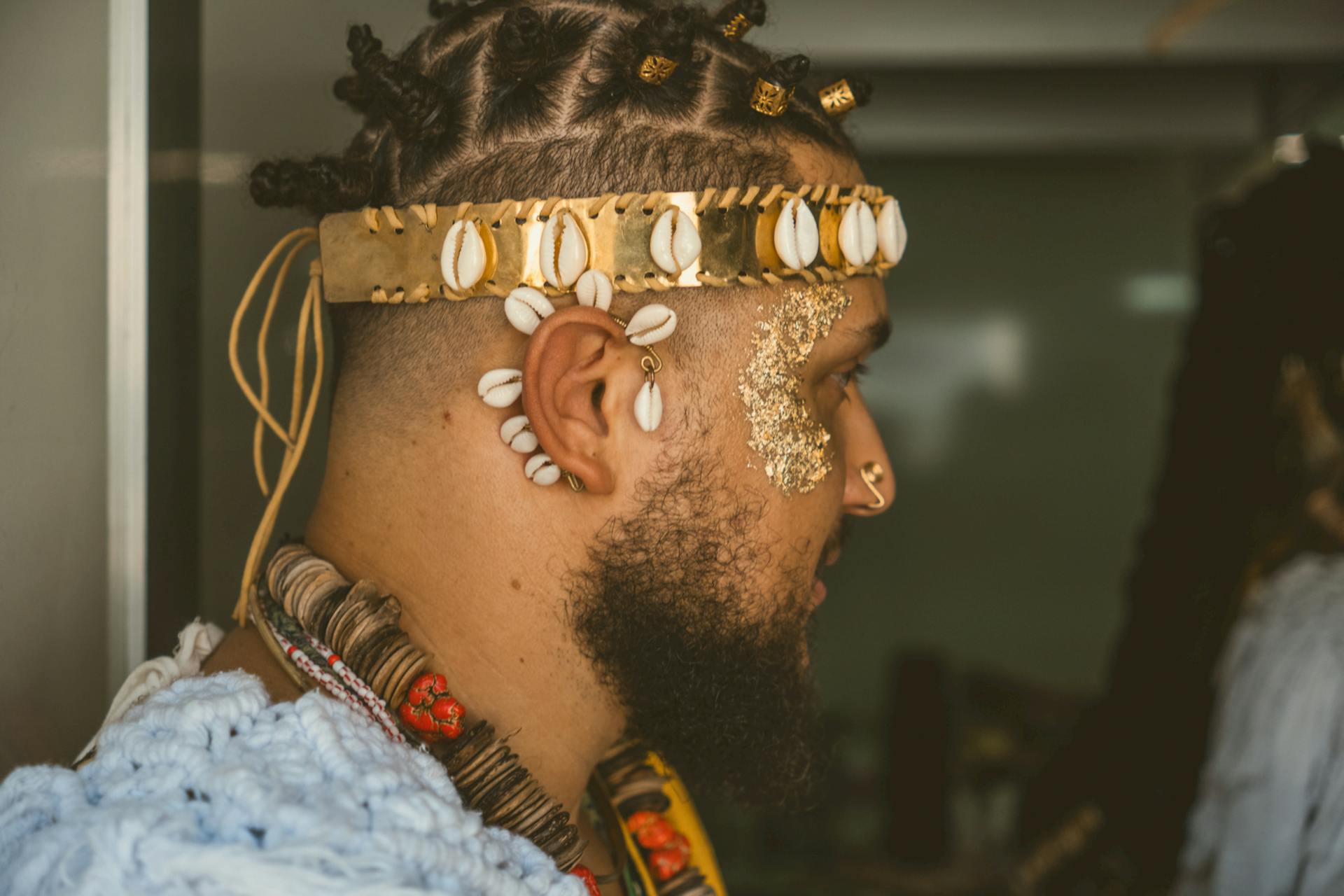 Side profile portrait of a man wearing traditional jewelry with cowrie shells and gold accents indoors.