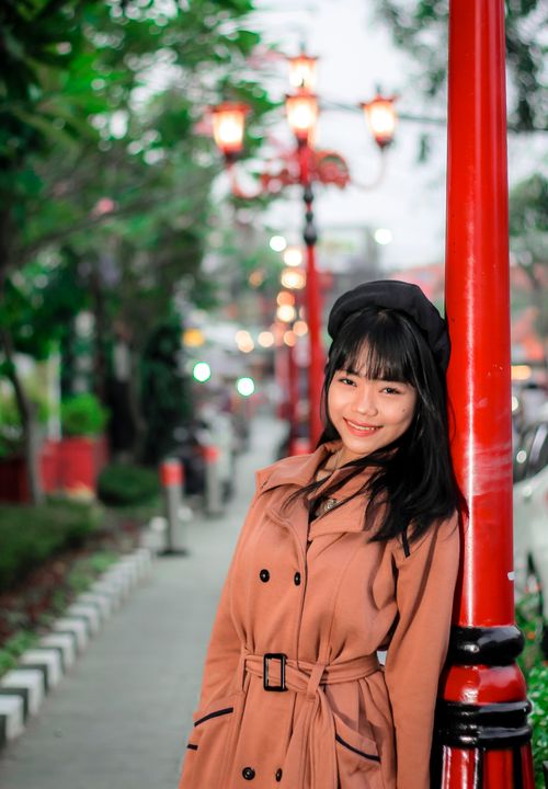 Free Smiling woman in a trench coat leans against a red lamp post in a vibrant urban street. Stock Photo