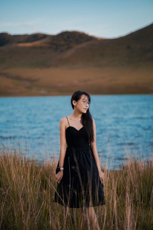 Free Young woman in a black dress stands by a tranquil lake with grassy hills in the background. Stock Photo