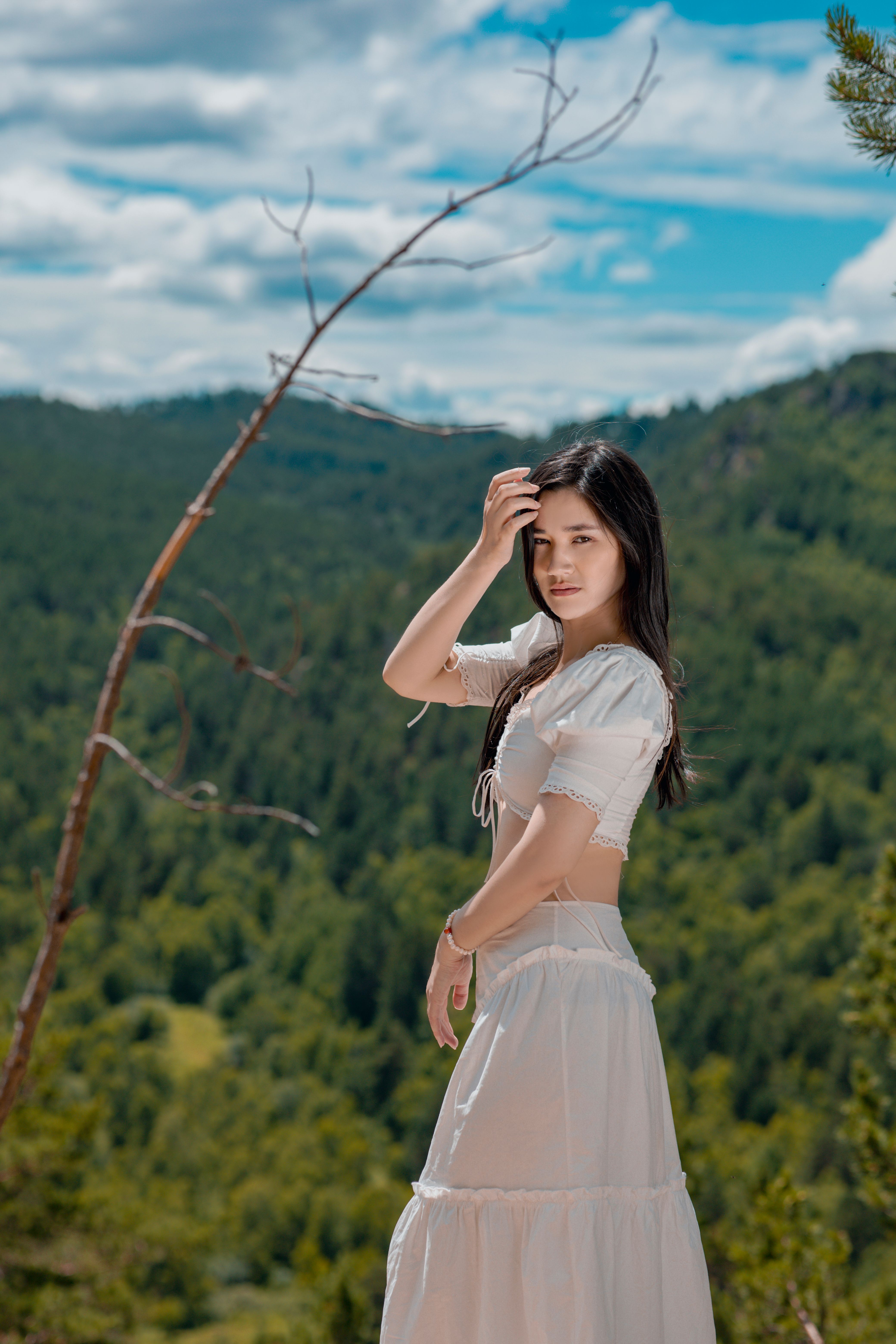 A young woman in a white dress poses outdoors in a lush summer forest.
