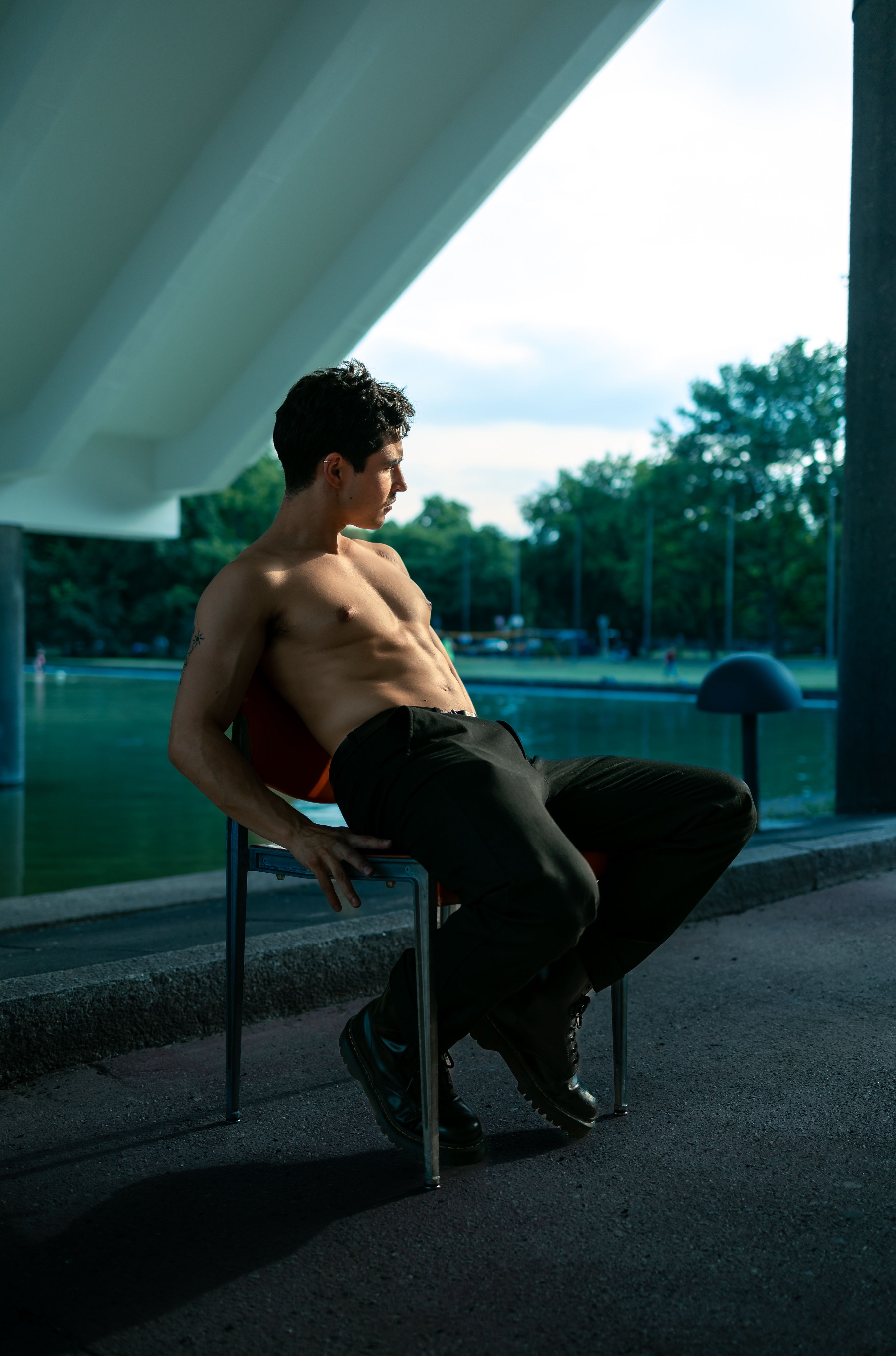 A shirtless man sits on a chair outdoors, under modern architecture, with a cool, vibrant mood.
