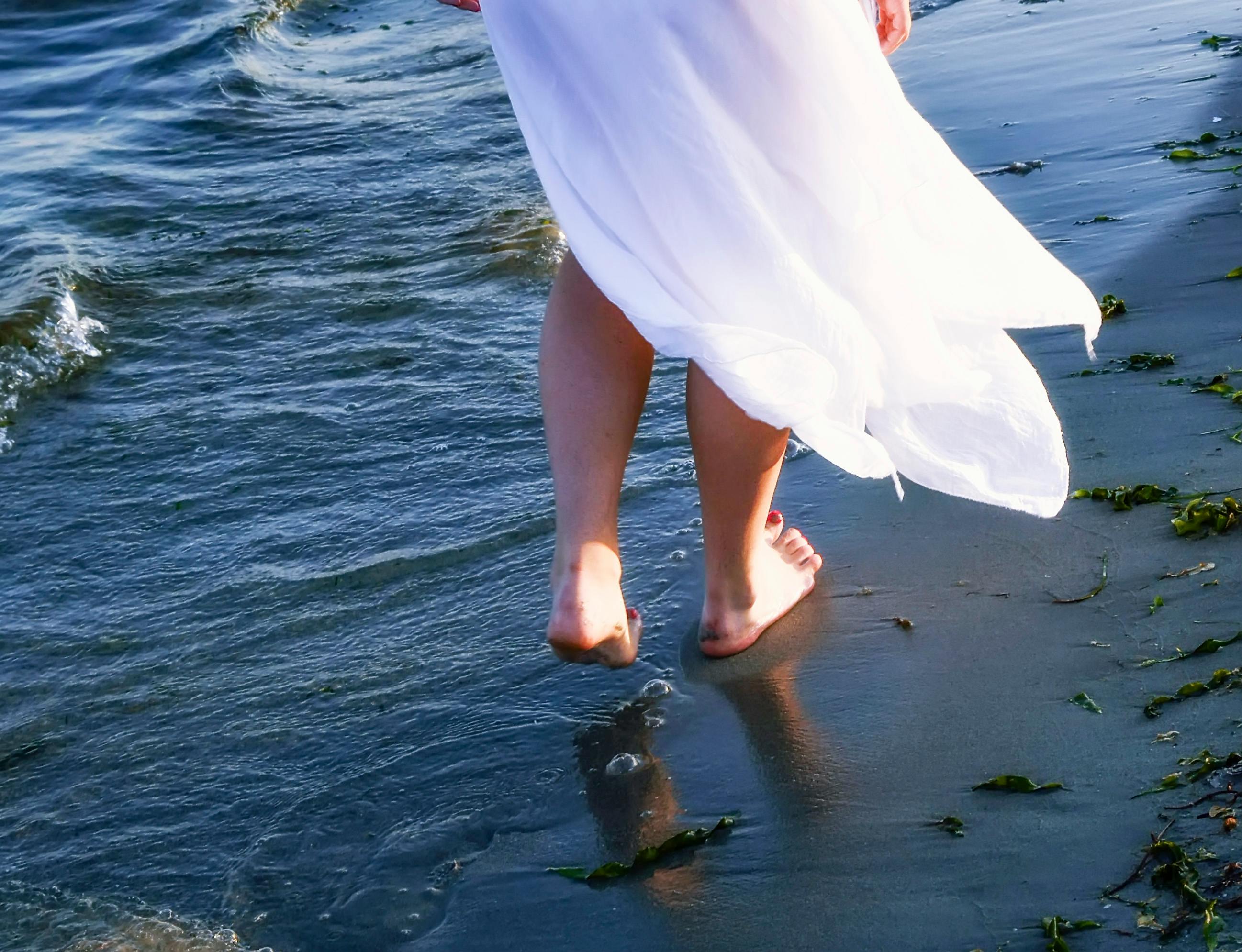A woman in a flowing white dress enjoys a peaceful walk along the beach shoreline.