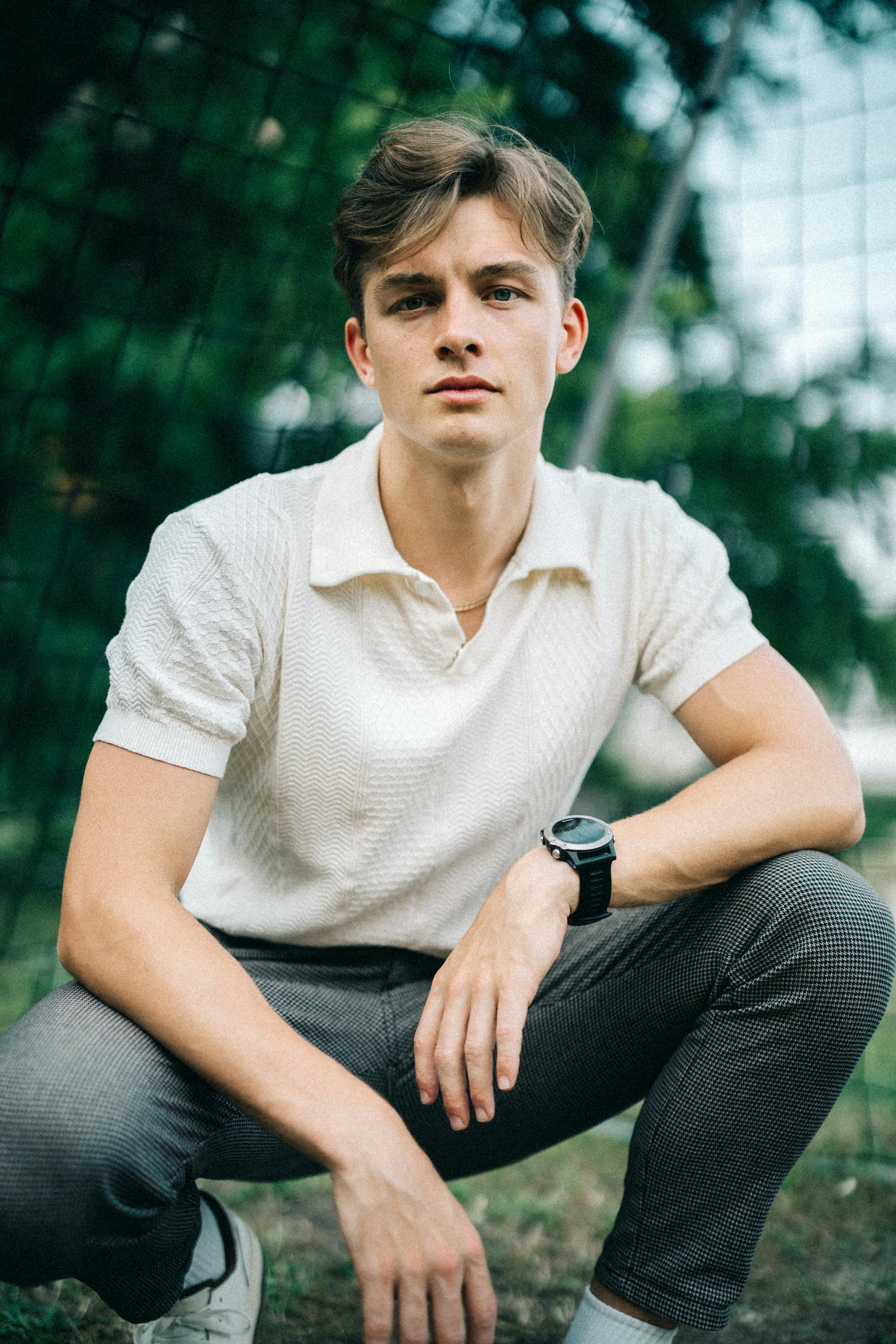 Young man crouching in a Berlin park, showcasing modern fashion and style on a sunny day.