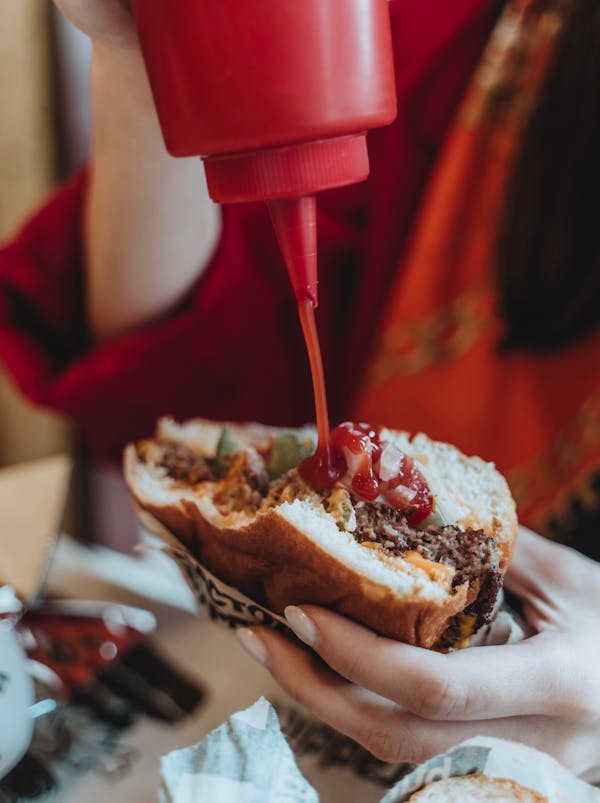 Crop faceless woman in red clothes adding ketchup to hamburger while eating in cafe