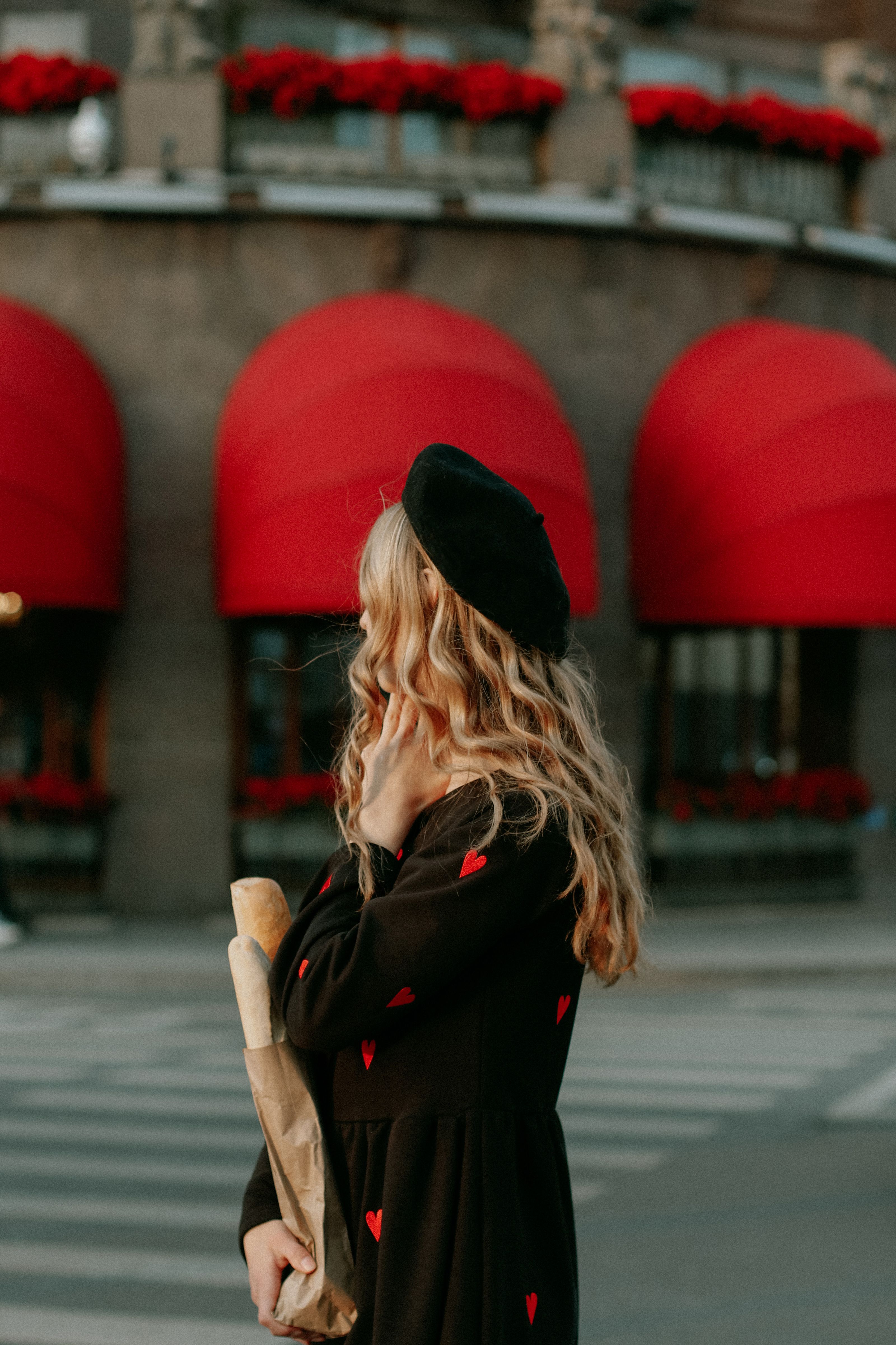 Mulher estilosa em vestido preto com corações vermelhos segurando uma baguete em uma rua com awnings vermelhos.