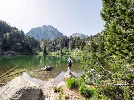 在西班牙的一个高山湖泊旁，一位徒步旅行者站在郁郁葱葱的绿植和风景秀丽的山丘之间