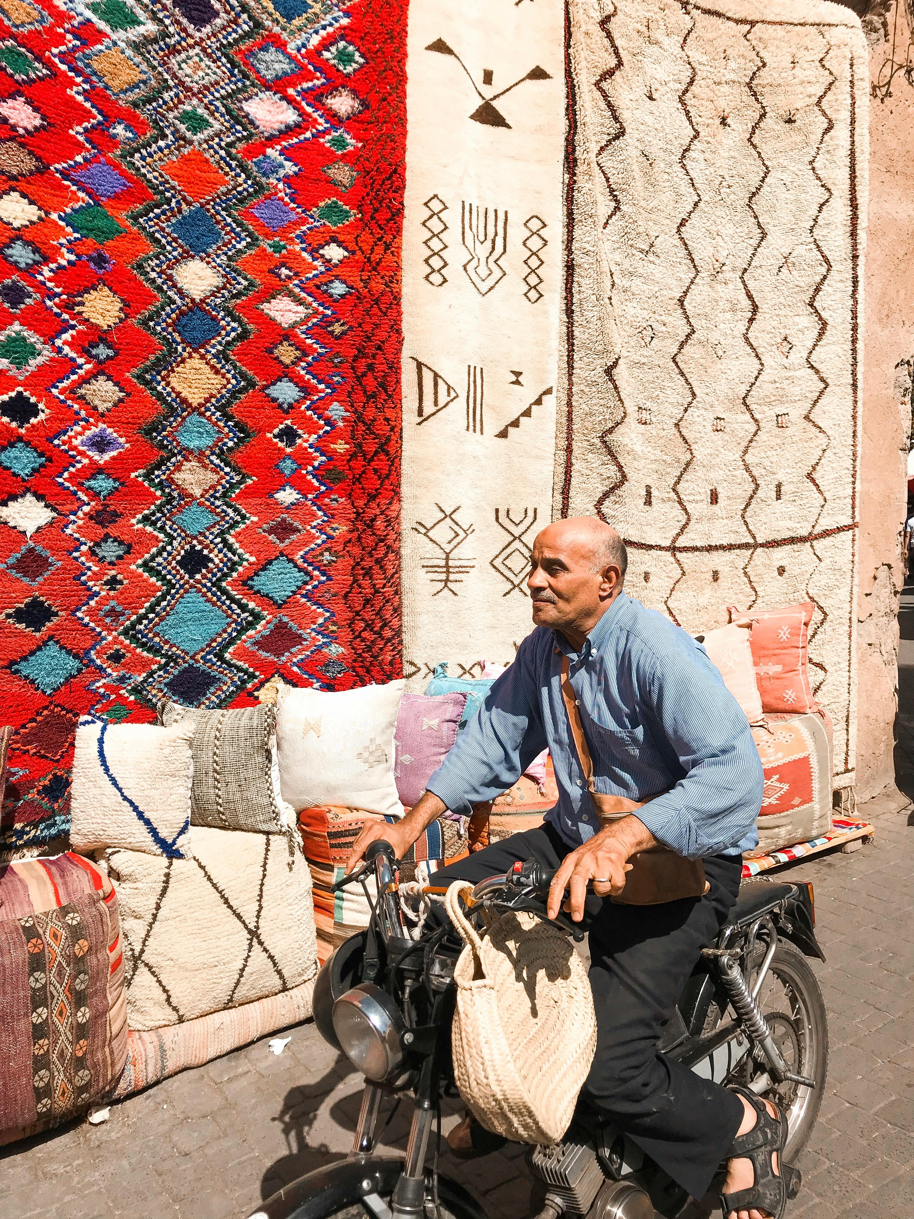 A man on a bike rides past vibrant rugs displayed in a sunny outdoor market.