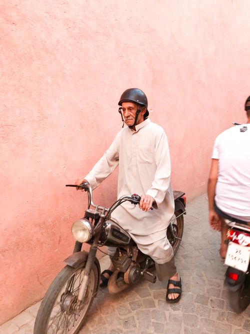 Free An elderly man rides a motorcycle down a narrow urban alley, wearing traditional attire and a helmet. Stock Photo